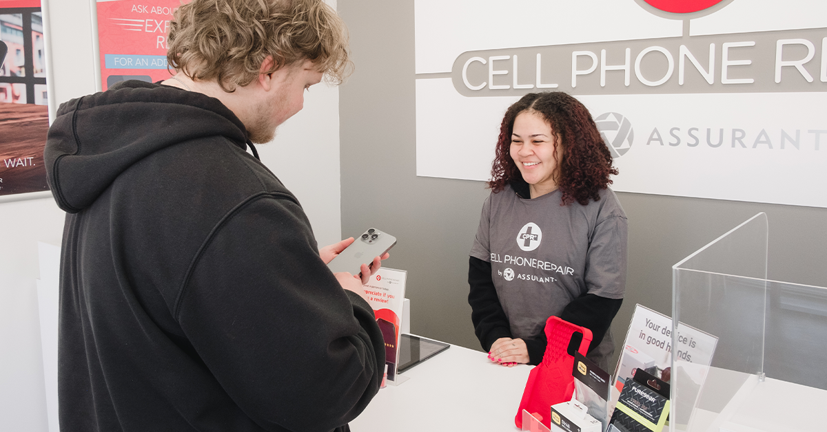 A consumer stands at the counter at an Assurant Authorized Repair Center location, preparing to hand their mobile device to a certified technician for their guaranteed repair appointment.