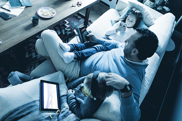 Dad seating on the couch with his kids, all three playing on electronic devices